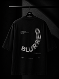 Premium black oversized T-shirt hanging on a wooden hanger featuring a minimalist white Japanese maple leaf print cascading from the right shoulder onto the chest. Studio product photography with a dark luxury background.