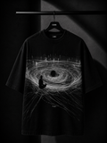 Black oversized T-shirt featuring a monochrome abstract vortex graphic with a meditating silhouette and flowing energy lines, displayed on a premium hanger in a luxury studio setting.