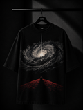 Black oversized T-shirt featuring a dramatic cosmic vortex graphic with a lone meditating silhouette on a red hill, displayed on a premium wooden hanger in a luxury dark studio setting.